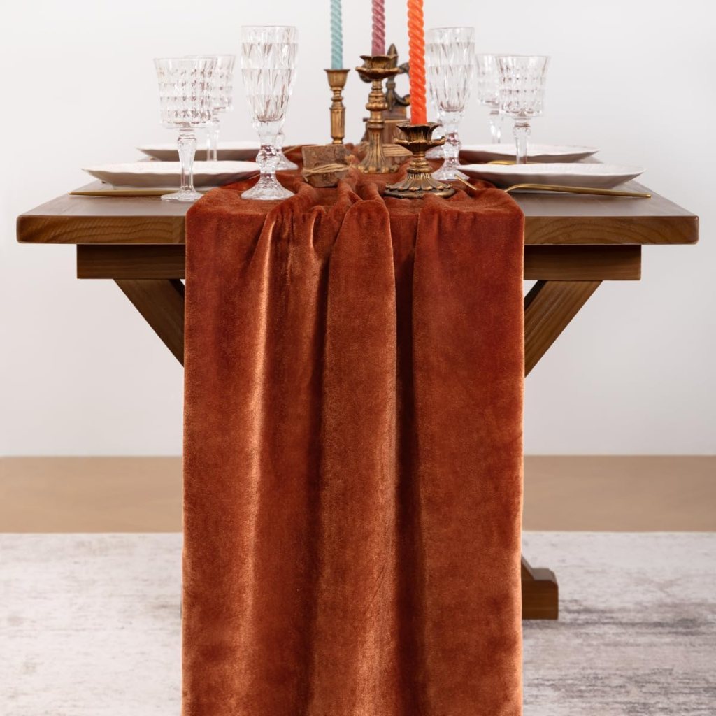 A wooden dining table set with a rich orange velvet table runner, elegant glassware, decorative candle holders, and plates arranged neatly.