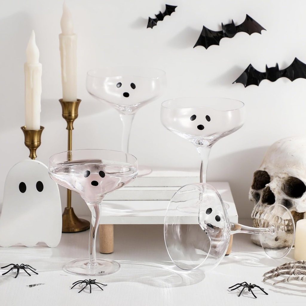 A festive Halloween-themed display featuring clear glasses with ghost faces, black bats, a skull, a white ghost decoration, candles, and black spiders.