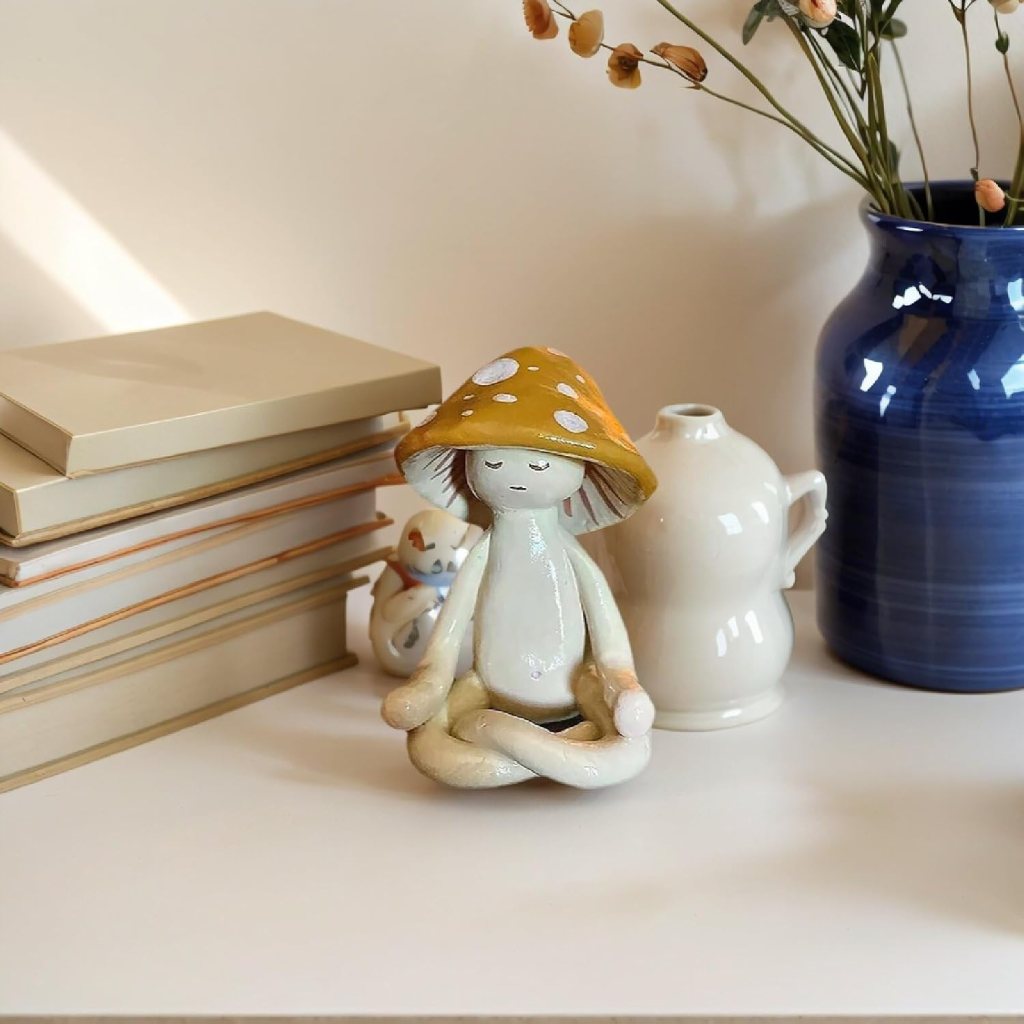 A whimsical ceramic figurine of a seated character wearing a yellow mushroom hat, surrounded by a stack of books, a white teapot, and a blue vase with dried flowers.