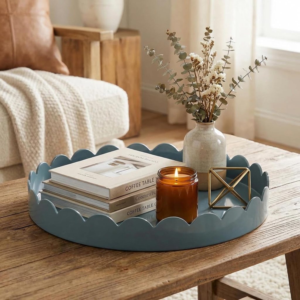 A blue scalloped tray on a wooden coffee table, holding stacked coffee table books, a lit candle in a brown jar, and a decorative golden geometric object, with a vase of dried flowers in the background.