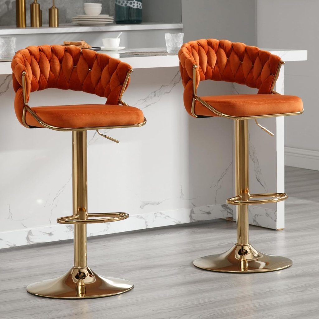 Two modern bar stools with orange quilted upholstery and gold metal bases, positioned at a marble countertop.