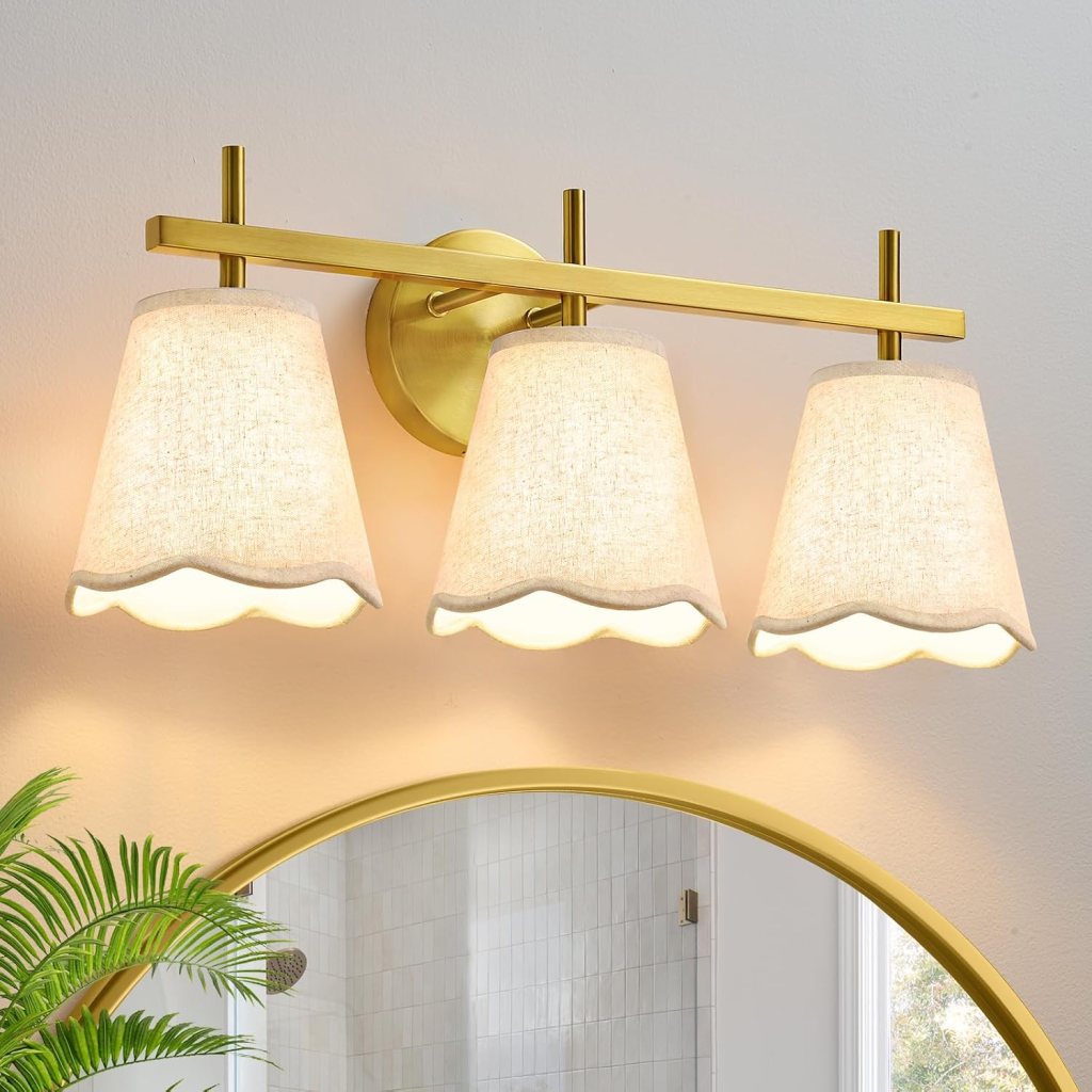 Three light fixtures with fabric shades mounted on a gold bar against a light wall, reflecting in a round mirror.