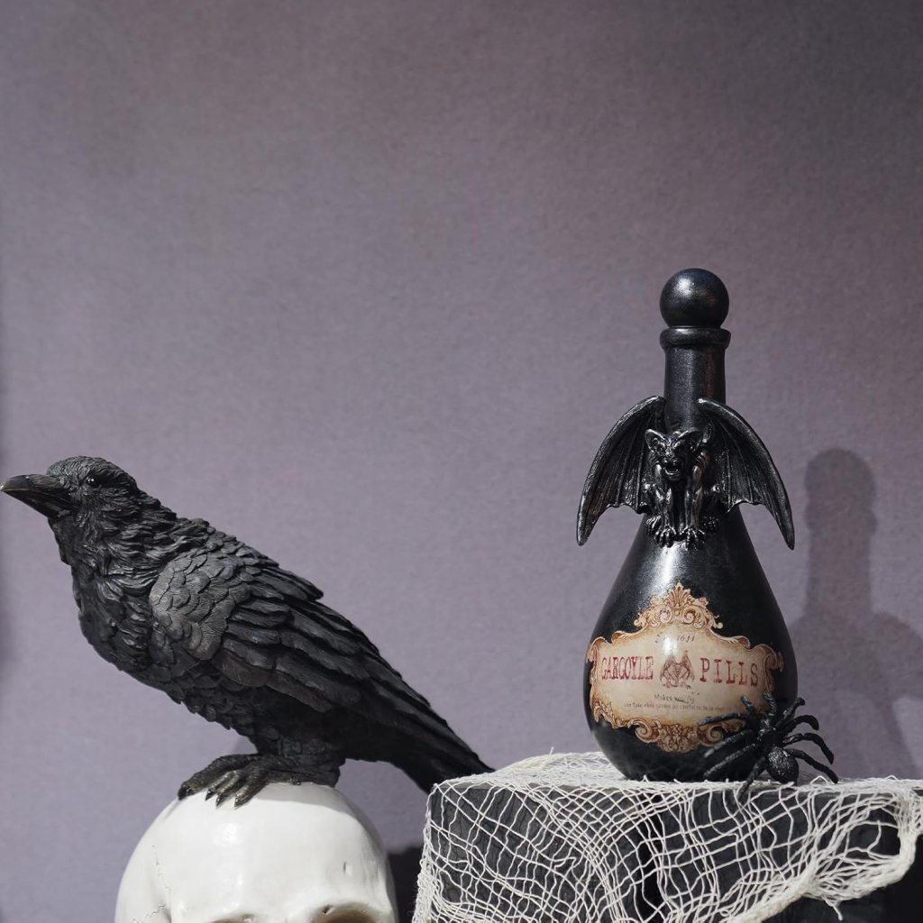 A decorative black crow perched on a white skull, next to an ornate black bottle labeled 'Gargoyle Pills' adorned with a bat figure on top and a spider nearby, all displayed on a netted background.