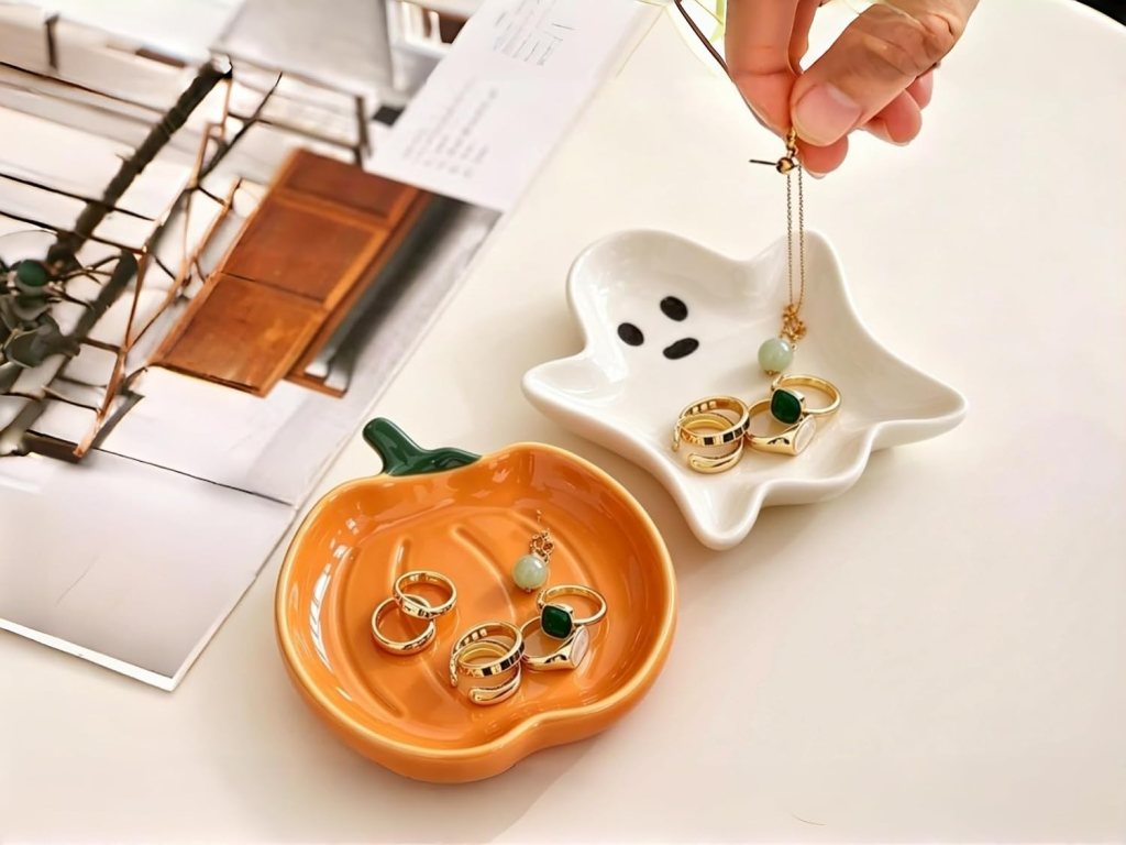 A hand holding a necklace above a white ghost-shaped dish, while various rings are arranged in an orange pumpkin-shaped dish and the background features a piece of paper.