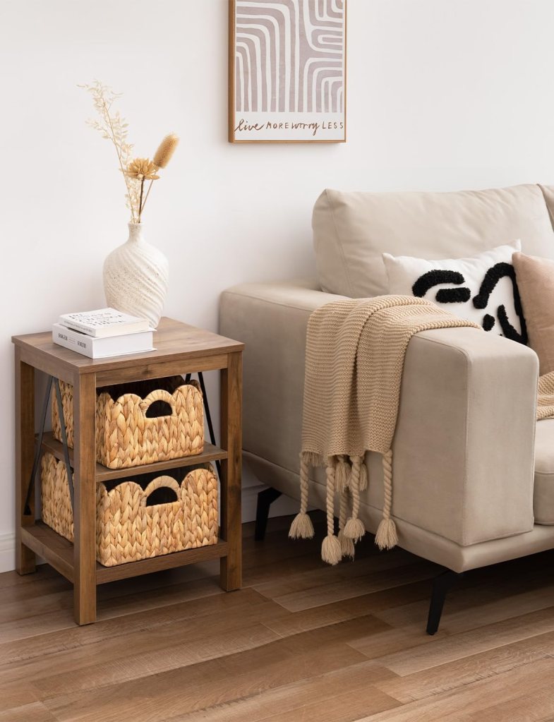 A modern living room setup featuring a beige sofa with decorative pillows, a knit throw blanket draped over the arm, a wooden side table with books, and two woven storage baskets underneath. A vase with dried flowers and an abstract framed wall art piece are also present.