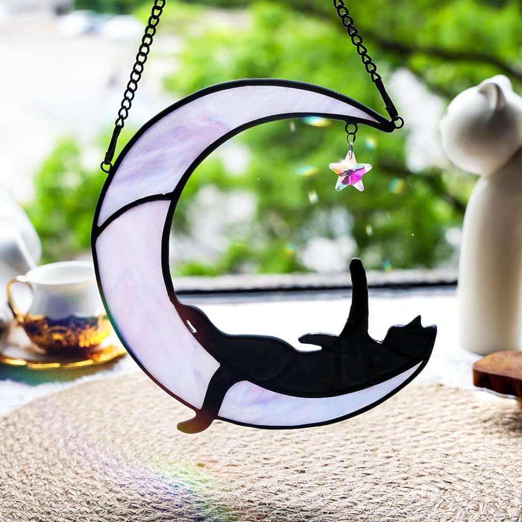 A stained glass decoration featuring a crescent moon with a black cat sleeping inside, suspended by a chain, with a star hanging from the moon.