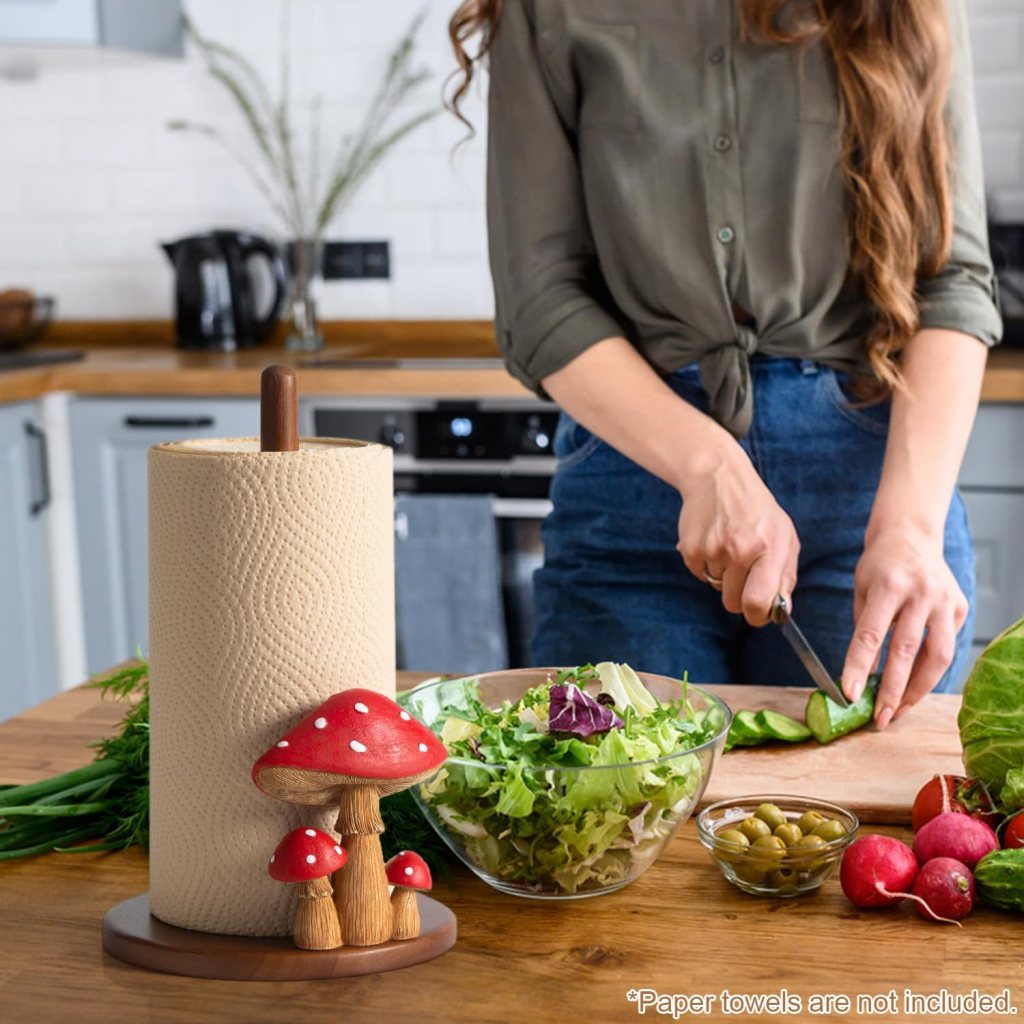 A woman chopping vegetables in a kitchen, with a decorative paper towel holder featuring mushrooms in the foreground and a bowl of salad nearby.