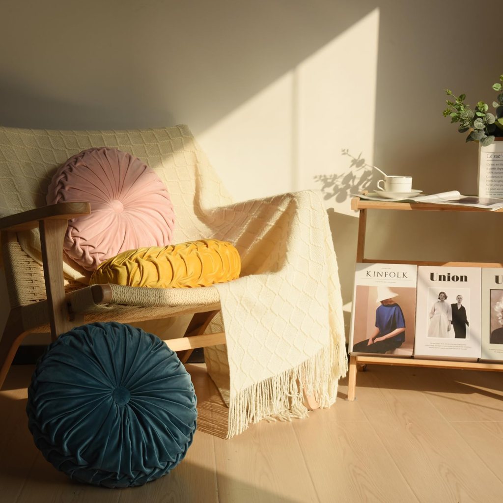 A cozy seating area featuring a wooden chair with a beige throw blanket, adorned with three colorful round pillows: pink, yellow, and teal. Nearby, a small wooden table displays magazines and a cup.