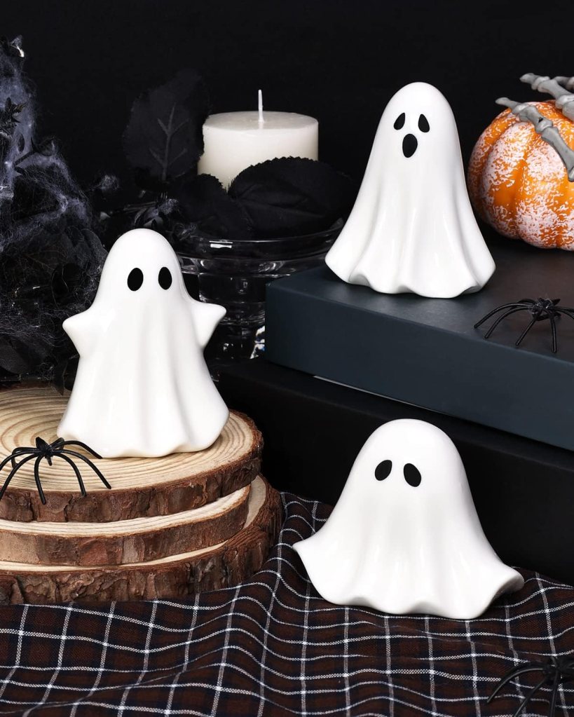 Three decorative white ghost figures with black eyes sitting on wooden slices, surrounded by a black cloth, a white candle, a pumpkin, and spider decorations.