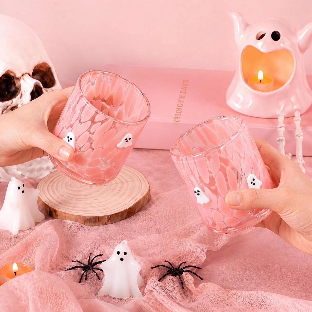 Two hands holding pink glasses with ghost designs, surrounded by white ghost candles, black spider decorations, and a pink candle in a ghost-shaped holder, on a pink tablecloth.