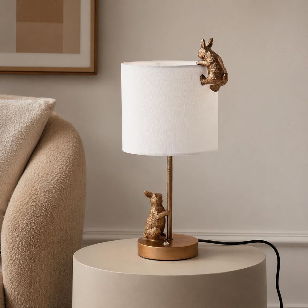 A decorative table lamp featuring two playful rabbit figures, one climbing on the lamp shade and the other sitting at the base, positioned on a minimalist round table.