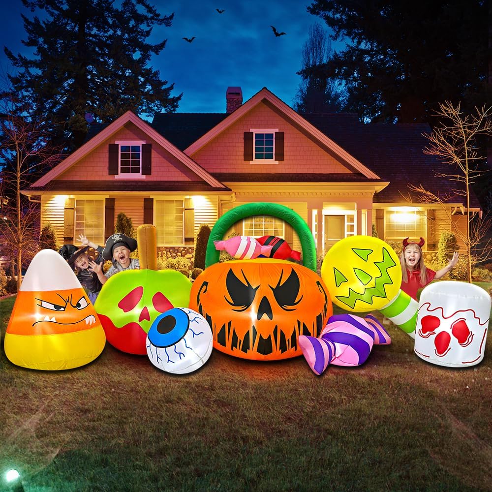 A festive Halloween scene featuring inflatable decorations including a large orange pumpkin with a spooky face, candy corn, and colorful lollipops. Two children are posing happily in the foreground, surrounded by a dark evening sky and a well-lit house.