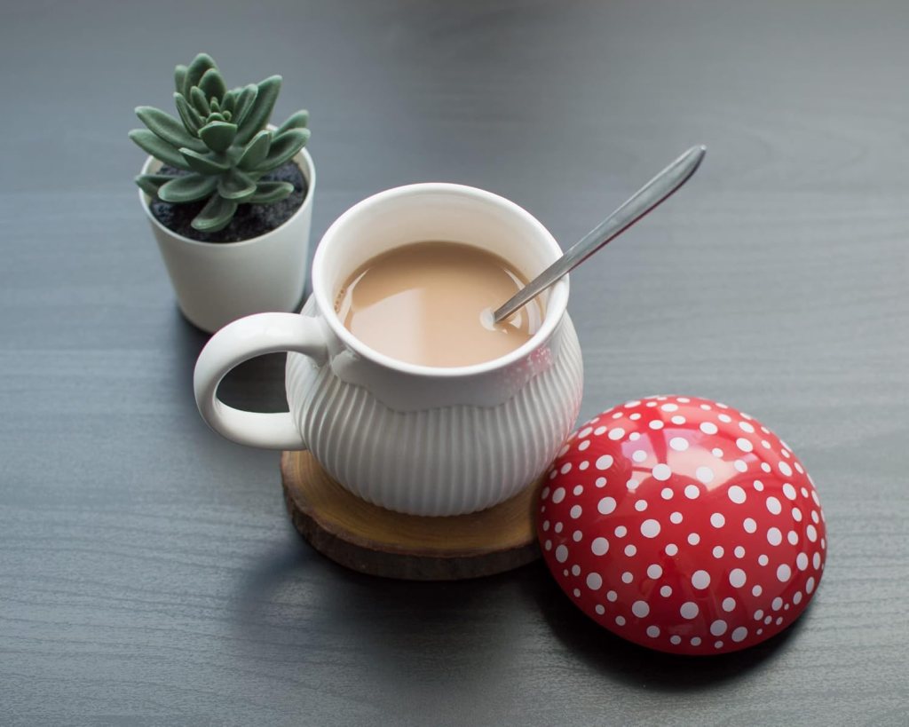 A white ceramic mug with brown tea or coffee and a spoon, placed on a wooden coaster beside a small potted succulent plant and a red polka-dotted lid.