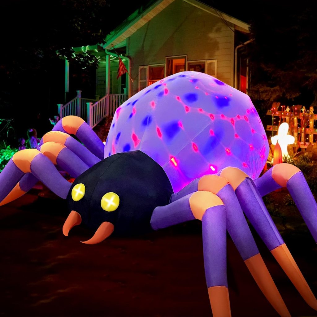 A large, inflatable spider with a glowing, colorful body and bright yellow eyes, set against a dark house backdrop decorated for Halloween.