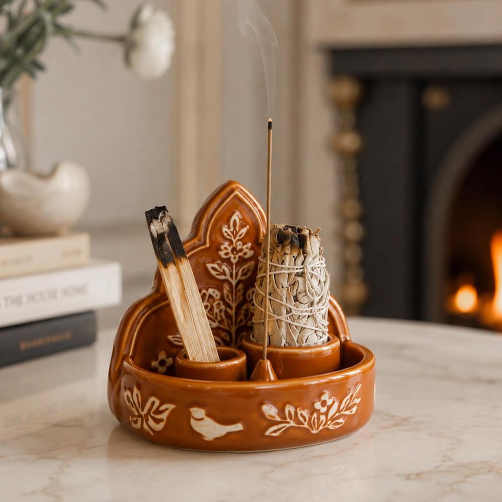 A decorative ceramic incense holder featuring a burnt stick of incense and a bundle of sage, set on a marble surface with a blurred fireplace in the background.