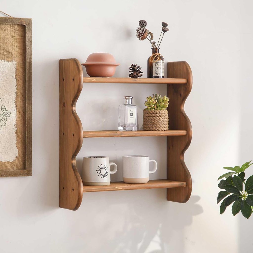 A wooden wall-mounted shelf with curved edges, displaying a pink bowl, a glass bottle, a potted succulent, two mugs, and decorative items including pine cones and dried flowers. A plant is visible beside the shelf.