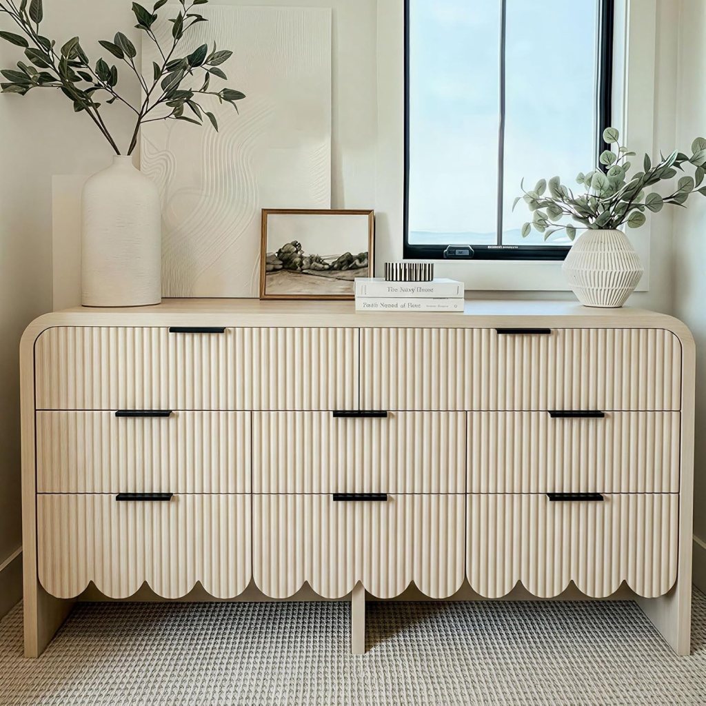 A modern wooden dresser with ribbed detailing and scalloped edges, topped with a decorative vase and a framed artwork, situated near a window in a cozy interior.