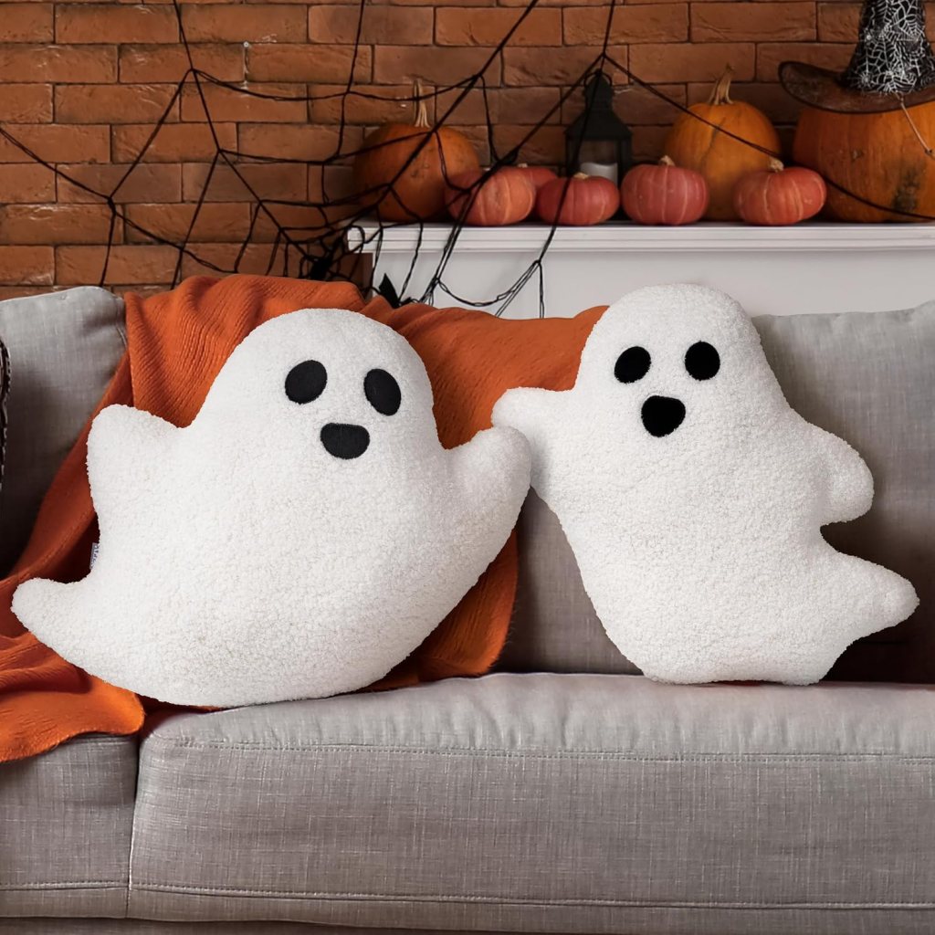 Two plush ghost-shaped pillows with black eyes and mouth, sitting on a gray sofa, draped with an orange blanket, with pumpkins and a spider web in the background.