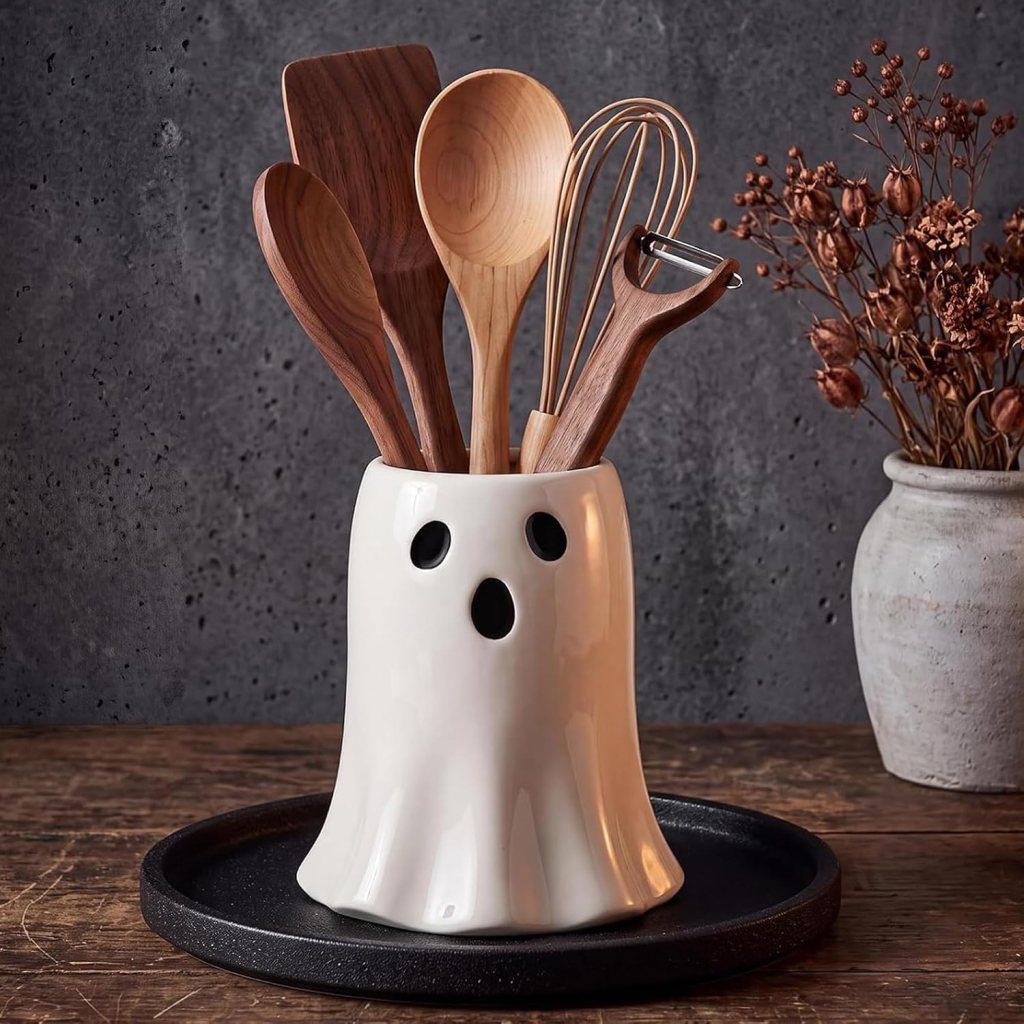 A ghost-shaped utensil holder with wooden kitchen tools inside, placed on a dark tray, with dried flowers in a vase nearby.