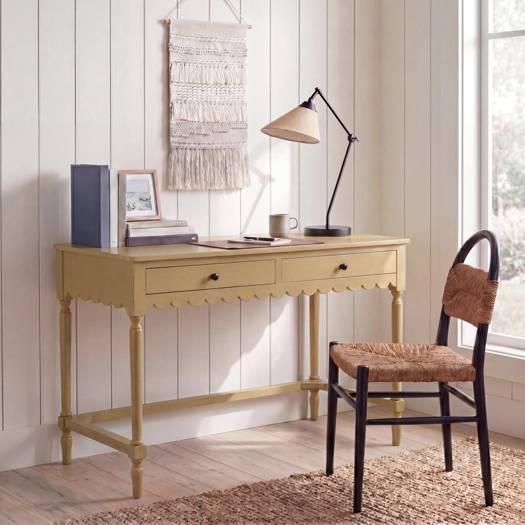A stylish yellow desk with two drawers, a desk lamp, and a chair with a woven seat, set against a light wooden wall and a large window.