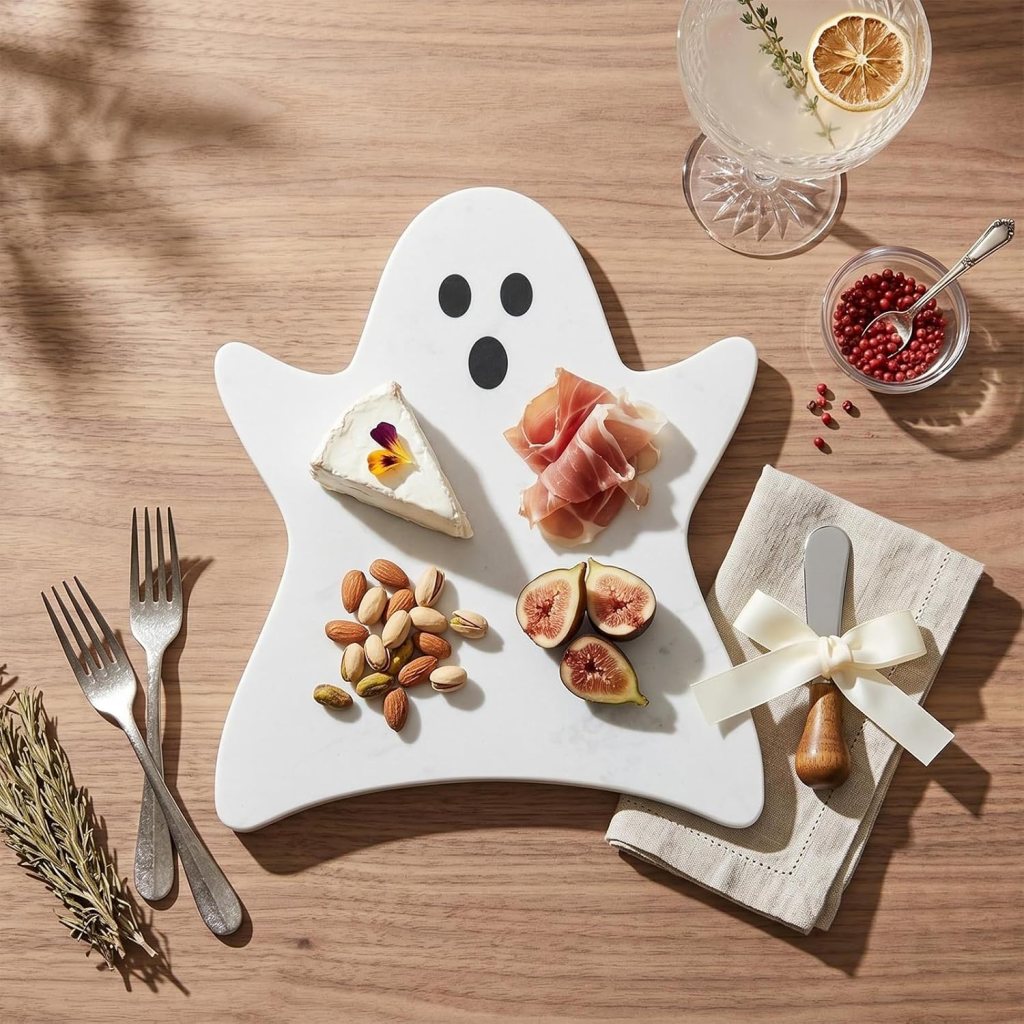 A ghost-shaped cheese board featuring a wedge of cheese with a flower, slices of prosciutto, fresh figs, and assorted nuts, accompanied by a glass of cocktail and a serving knife on a wooden table.
