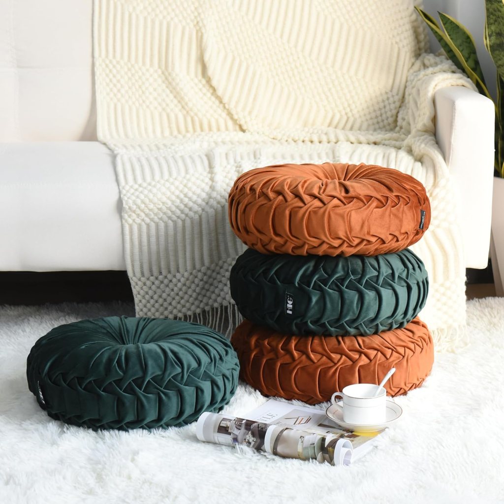 A cozy living space featuring four decorative round pillows in earthy colors (green and orange) stacked beside a white sofa, with a textured blanket draped over it. A small white teacup and magazines are placed on the floor.