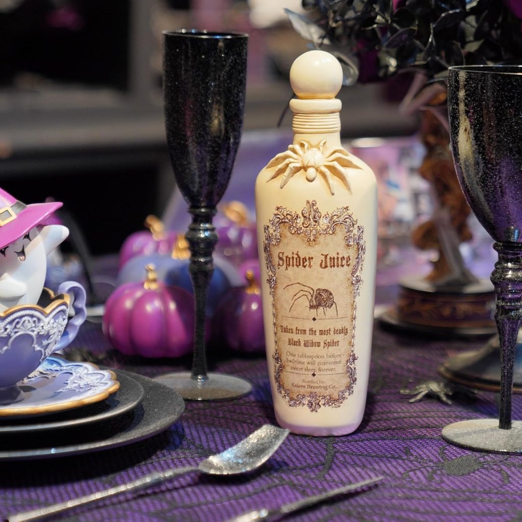 A decorative bottle labeled 'Spider Juice' with a spider illustration, displayed on a Halloween-themed table setting with purple and black decor, surrounded by elegant dishware and small purple pumpkins.