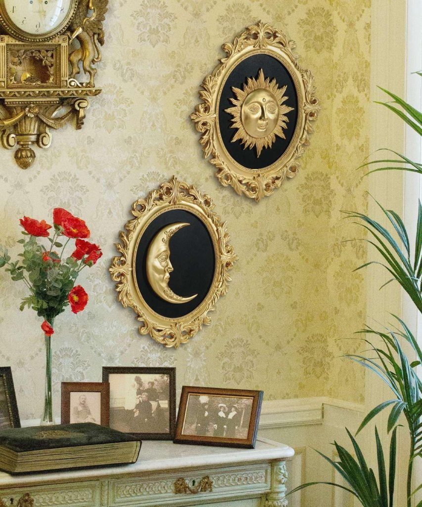 A decorative wall featuring two framed gold sun and moon face artworks against a patterned wallpaper, alongside a vase with red flowers and various framed photographs on a table.