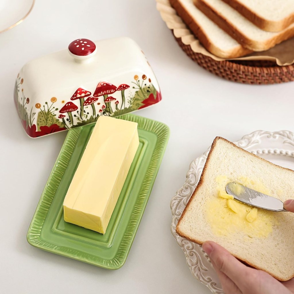 A block of butter on a green serving dish beside a decorative butter dish with mushrooms, with a hand spreading butter on a slice of bread on an elegant plate.