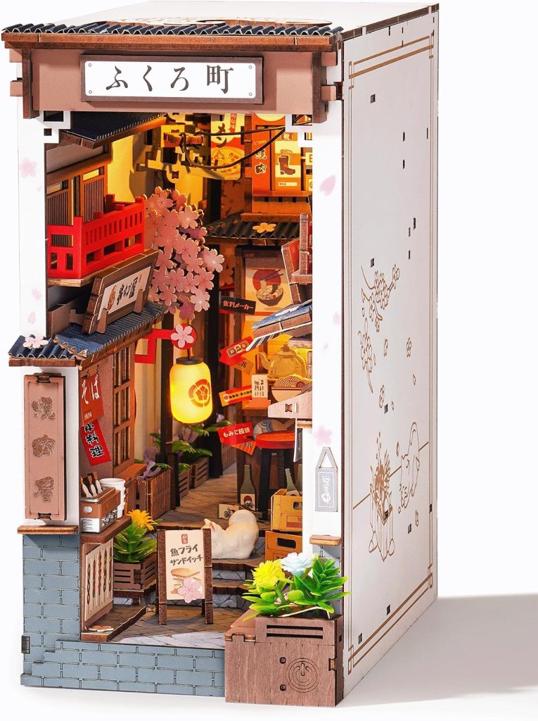 A detailed miniature model of a traditional Japanese street scene, featuring storefronts, lanterns, cherry blossom decorations, and small potted plants.