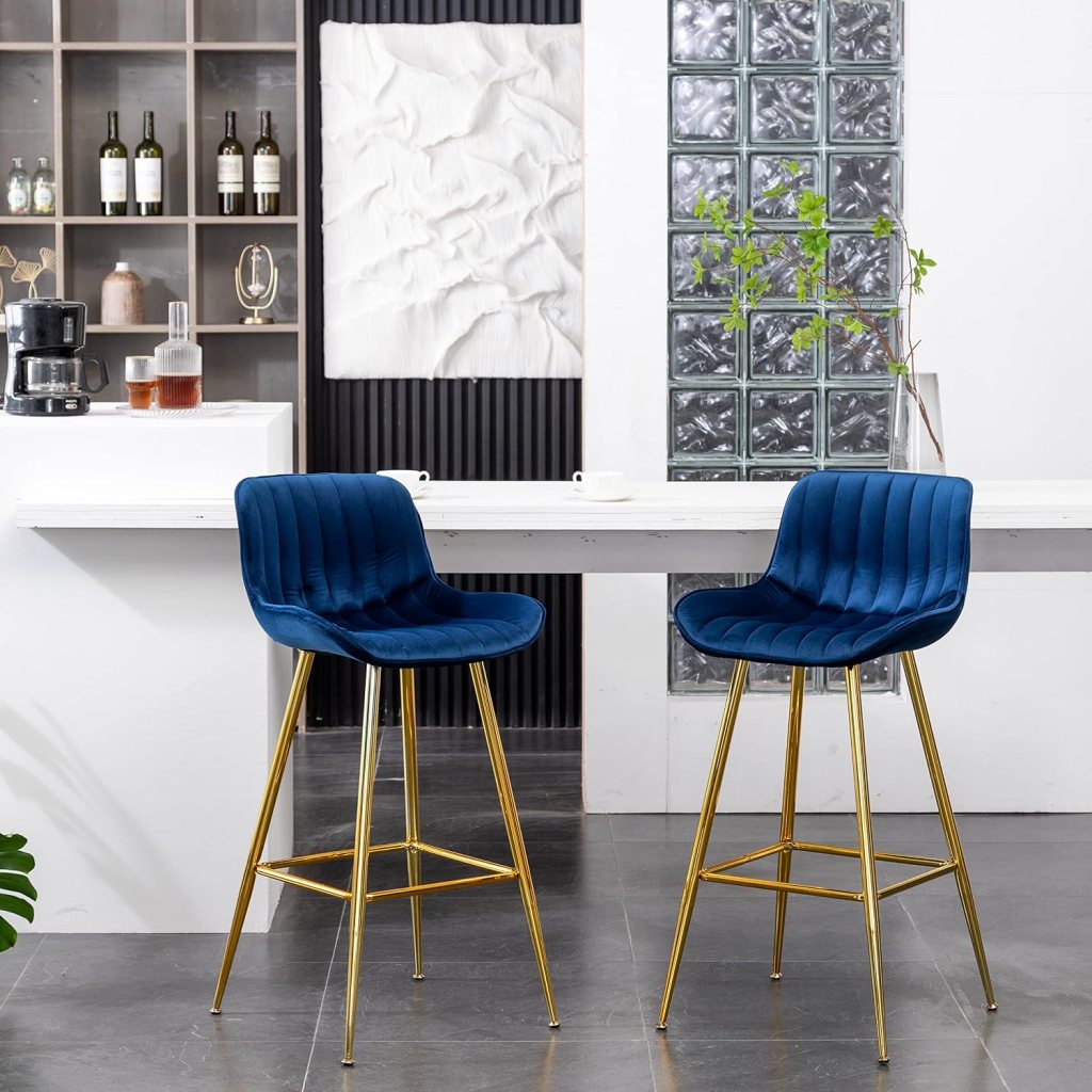 Two blue velvet bar stools with gold legs are positioned at a white wooden countertop in a modern kitchen setting, featuring a shelf with wine bottles and a decorative wall art piece in the background.
