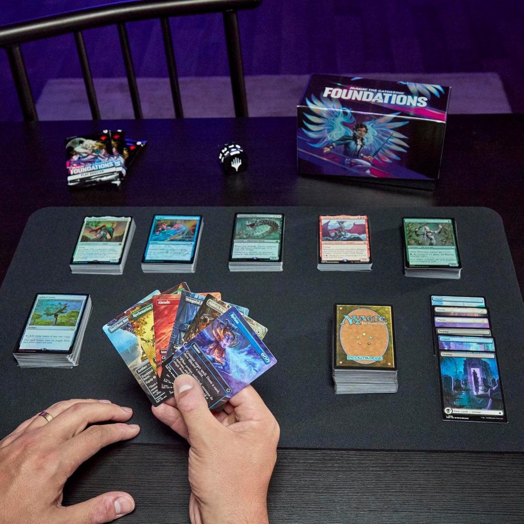 A person holding several Magic: The Gathering cards with a game setup on a black table, including a box labeled 'FOUNDATIONS', various cards arranged in rows, and game dice.