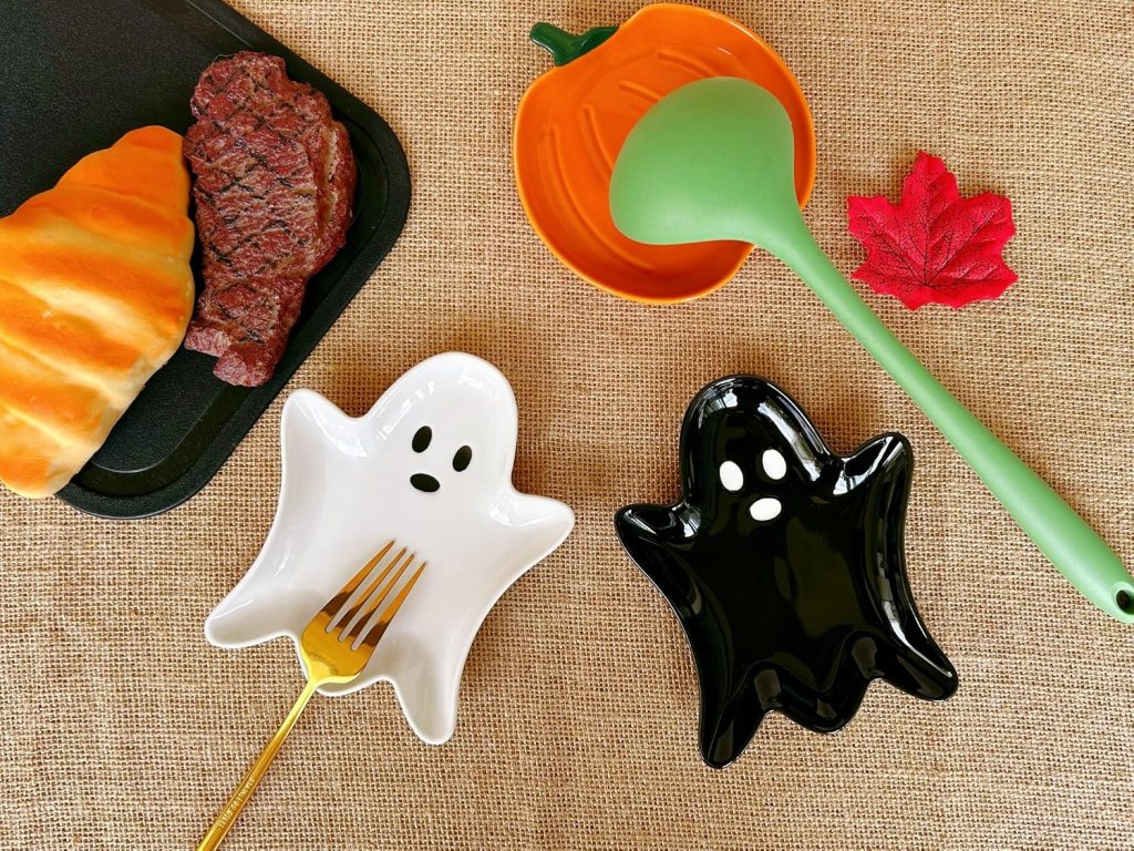 An overhead view of a Halloween-themed dining setup featuring ghost-shaped dishes, a golden fork, a carved pumpkin dish, a green ladle, colorful autumn leaves, and a steak alongside a bread roll on a black tray.