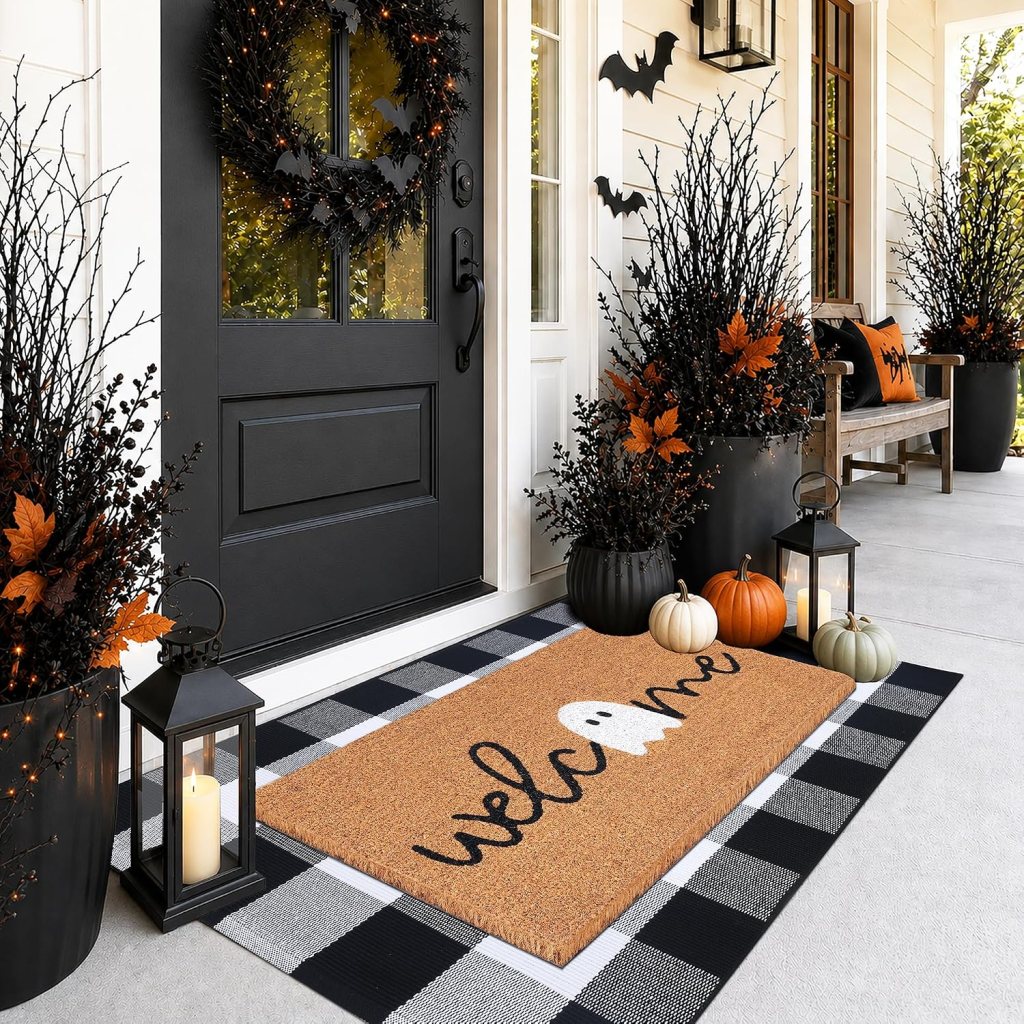 A stylish front porch decorated for fall and Halloween, featuring a black door, a witch-themed wreath, and a welcome mat with a ghost design. There are decorative pumpkins, tall black planters with branches and bat accents, and lanterns adding ambiance.