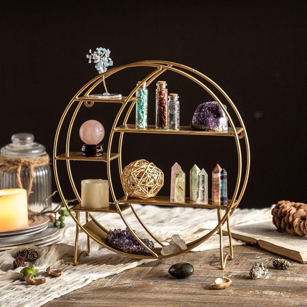 A decorative gold metal display shelf featuring various crystals, gemstones, and small jars, set against a dark background. Items include a rose quartz sphere, amethyst clusters, and a small wire tree, with candles and natural elements in the foreground.