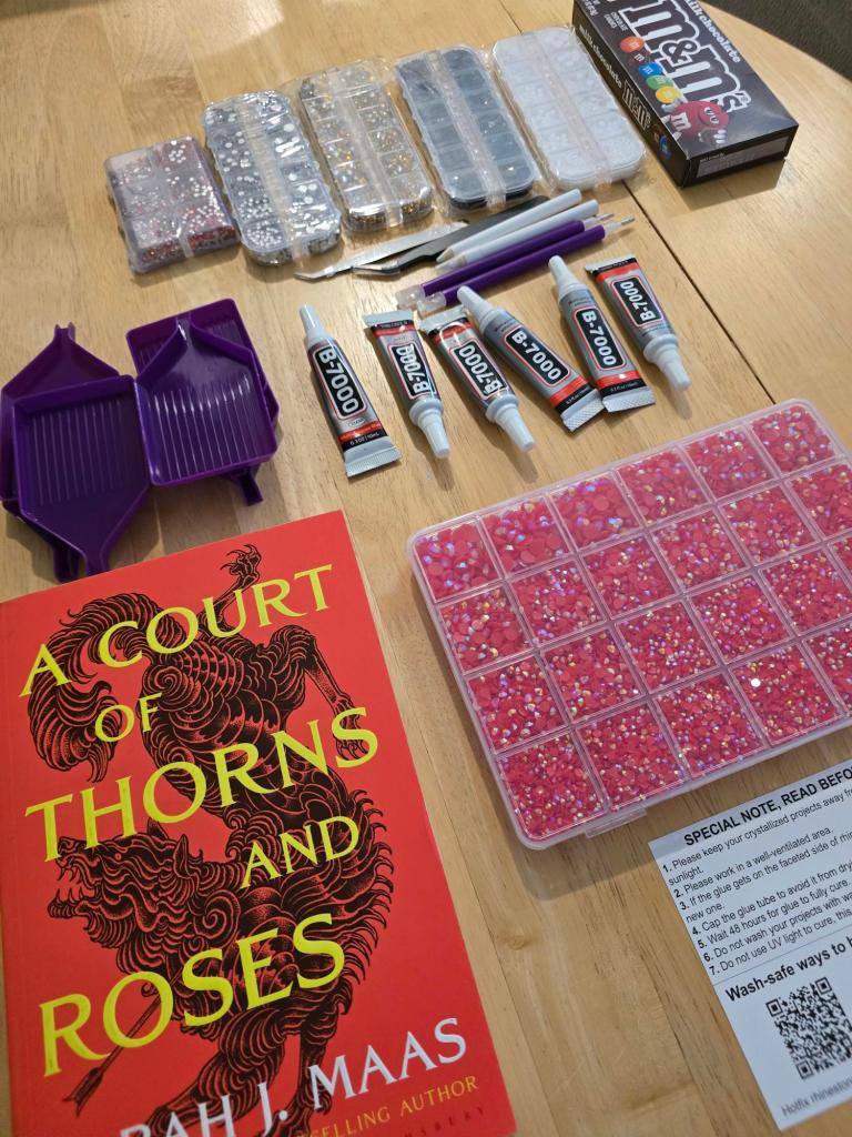 A flat lay of crafting supplies including two purple trays, several containers of colorful rhinestones, adhesive glue tubes, a pair of tweezers, and a book titled 'A Court of Thorns and Roses' by Sarah J. Maas on a wooden surface.