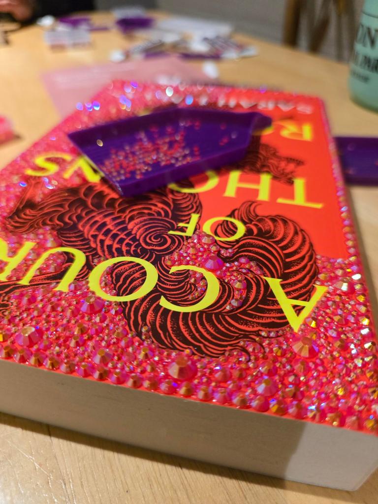 A colorful book titled 'A Court of Thorns and Roses' with a glittering pink and red cover, adorned with rhinestones, placed on a wooden table, alongside purple crafting supplies.